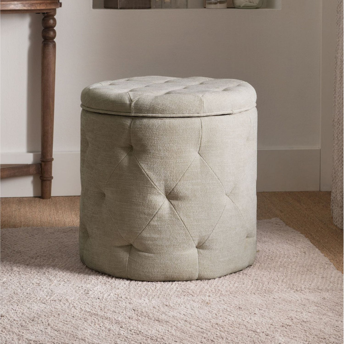 Beige tufted storage ottoman in a room with wooden floor and wall.